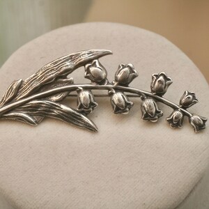 May include: A silver-toned brooch in the shape of a lily of the valley plant. The brooch features detailed leaves and bell-shaped flowers. The design is set against a neutral background, highlighting the intricate metalwork.