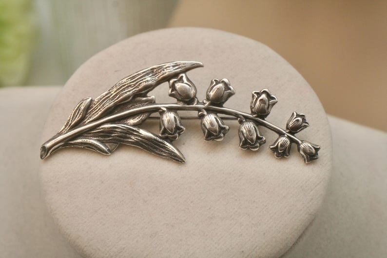 May include: A silver-toned brooch in the shape of a lily of the valley plant. The brooch features detailed leaves and bell-shaped flowers. The design is set against a neutral background, highlighting the intricate metalwork.