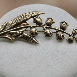 May include: A bronze-toned metal brooch shaped like a lily of the valley flower. The design features a long, textured leaf extending to the left and a curved stem on the right adorned with seven small, bell-shaped flower blossoms in a row.