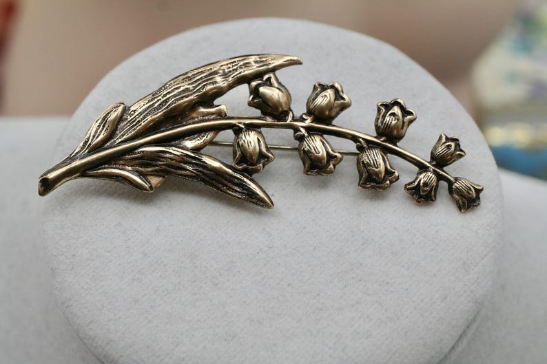 May include: A bronze-toned metal brooch shaped like a lily of the valley flower. The design features a long, textured leaf extending to the left and a curved stem on the right adorned with seven small, bell-shaped flower blossoms in a row.