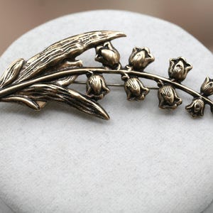 May include: A bronze-toned metal brooch shaped like a lily of the valley flower. The design features a textured leaf on the left side and a curved stem extending to the right, adorned with seven small, bell-shaped flower blossoms in a row.