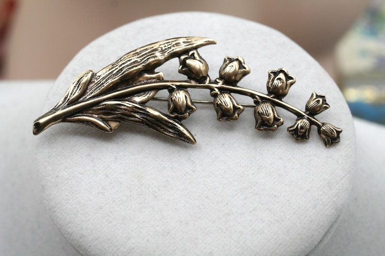 May include: A bronze-toned metal brooch shaped like a lily of the valley flower. The design features a textured leaf on the left side and a curved stem extending to the right, adorned with seven small, bell-shaped flower blossoms in a row.