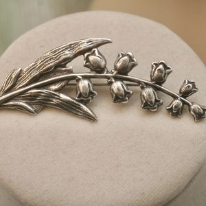 May include: A silver-toned brooch in the shape of a lily of the valley. The brooch features detailed leaves and bell-shaped flowers. The design is a classic floral motif, suitable for adding a touch of elegance to any outfit.