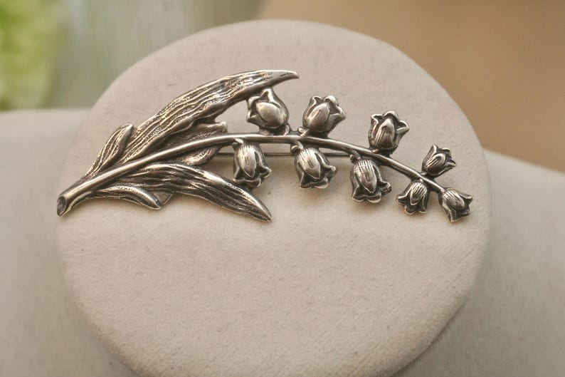 May include: A silver-toned brooch in the shape of a lily of the valley. The brooch features detailed leaves and bell-shaped flowers. The design is a classic floral motif, suitable for adding a touch of elegance to any outfit.