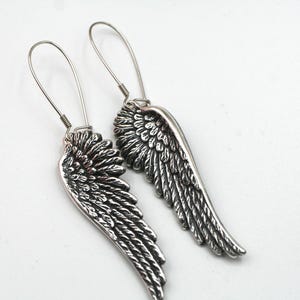 May include: A pair of silver-toned wing earrings. Each earring features a detailed wing design with intricate feather detailing. The earrings have a hook closure and are set against a white background.