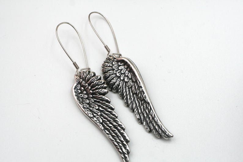 May include: A pair of silver-toned wing earrings. Each earring features a detailed wing design with intricate feather detailing. The earrings have a hook closure and are set against a white background.