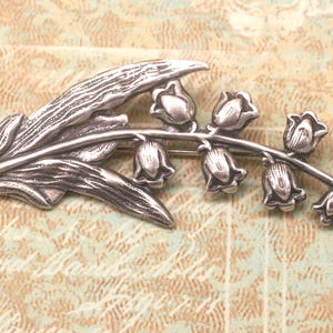 May include: A silver-toned brooch in the shape of a lily of the valley. The brooch features a detailed leaf and a stem with several bell-shaped flowers. The metal has a slightly aged appearance, giving it a vintage feel. The brooch is a decorative accessory.