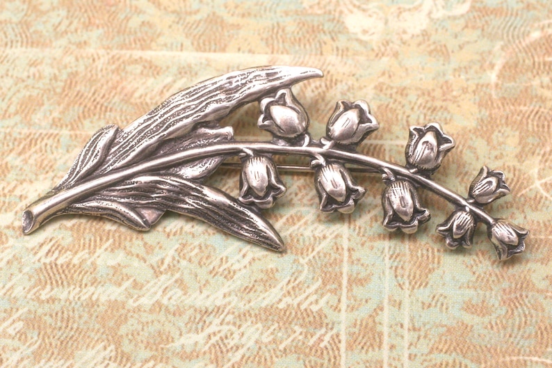 May include: A silver-toned brooch in the shape of a lily of the valley. The brooch features a detailed leaf and a stem with several bell-shaped flowers. The metal has a slightly aged appearance, giving it a vintage feel. The brooch is a decorative accessory.
