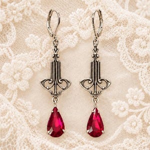 May include: A pair of silver-toned drop earrings featuring an intricate metalwork design with a vertical structure and a teardrop-shaped red gemstone pendant suspended from the base of each earring, secured with a lever-back closure.