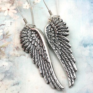 May include: A pair of silver-colored wing earrings. Each earring features a detailed feather design, with a curved shape. The earrings are suspended from a simple wire hook. The background has a soft, floral pattern.
