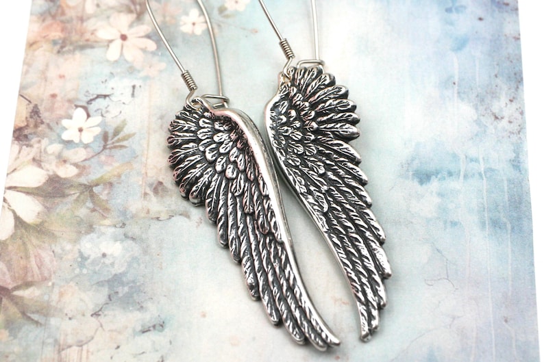 May include: A pair of silver-colored wing earrings. Each earring features a detailed feather design, with a curved shape. The earrings are suspended from a simple wire hook. The background has a soft, floral pattern.