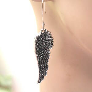 May include: A single silver-colored wing earring. The earring is a detailed representation of a bird's wing, with individual feather detailing. The earring hangs from a simple hook. The background is blurred, with a soft, neutral color palette.