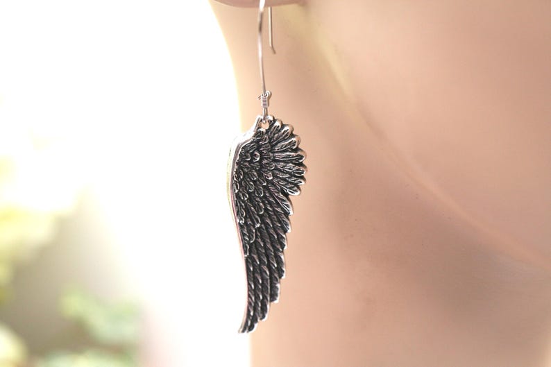 May include: A single silver-colored wing earring. The earring is a detailed representation of a bird's wing, with individual feather detailing. The earring hangs from a simple hook. The background is blurred, with a soft, neutral color palette.
