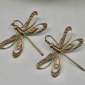 Può includere: Un paio di orecchini a forma di libellula dorati con dettagli intricati. Gli orecchini sono in metallo e hanno un design delicato e femminile.