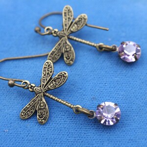 May include: A pair of dragonfly earrings. The earrings feature detailed bronze-colored dragonfly charms with dangling, round, light purple crystals. The earrings are displayed on a blue surface.