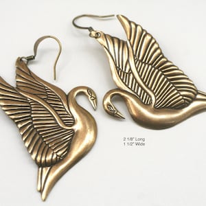 May include: A pair of bronze-colored swan earrings. Each earring features a swan with outstretched wings, detailed with feather textures. The earrings are 2 1/8 inches long and 1 1/2 inches wide.