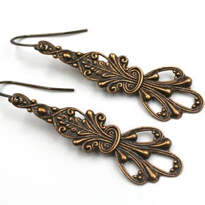 May include: A pair of bronze-colored filigree earrings. Each earring features an ornate design with floral and scrollwork details, including a central knot and teardrop-shaped elements. The earrings have a hook closure.