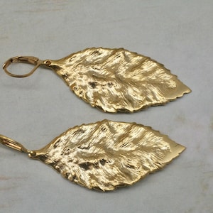 Könnte beinhalten: Ein Paar goldene Ohrringe in Blattform mit einer strukturierten Oberfläche. Die Ohrringe haben einen einfachen Hakenverschluss.