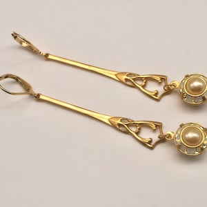 May include: A pair of gold-toned drop earrings. Each earring features a long, slender bar with an ornate design near the top and a round, pearl-like bead at the bottom. The earrings are set against a light background.
