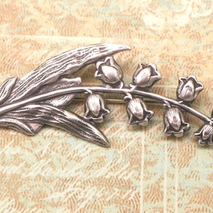 May include: Silver-toned brooch in the shape of a lily of the valley. The brooch features a detailed leaf and a stem with several bell-shaped flowers. The design is set against a textured, light brown background.