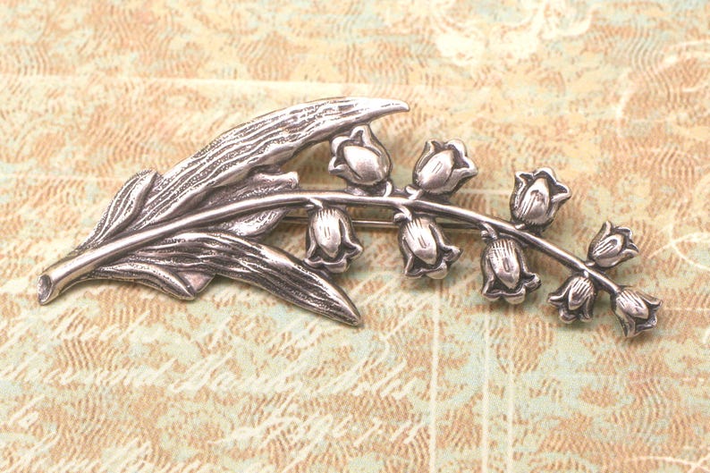 May include: Silver-toned brooch in the shape of a lily of the valley. The brooch features a detailed leaf and a stem with several bell-shaped flowers. The design is set against a textured, light brown background.
