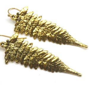 May include: A pair of gold-colored fern leaf earrings. The earrings are detailed with textured surfaces, and the hooks are visible. The earrings are set against a white background, highlighting the intricate design.