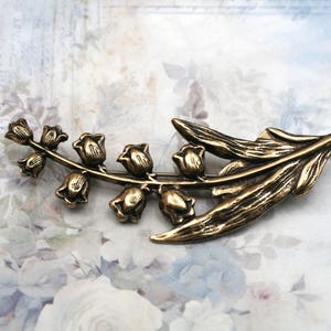 May include: A bronze-toned metal brooch shaped like a lily of the valley flower. The piece features a curved stem with six small bell-shaped blossoms and two elongated, textured leaves extending from the base, showcasing a metallic finish.