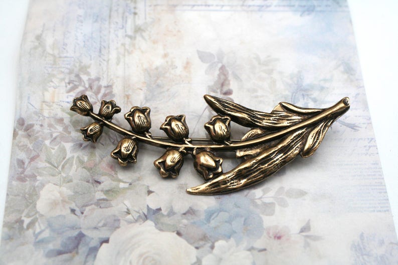 May include: A bronze-toned metal brooch shaped like a lily of the valley flower. The piece features a curved stem with six small bell-shaped blossoms and two elongated, textured leaves extending from the base, showcasing a metallic finish.