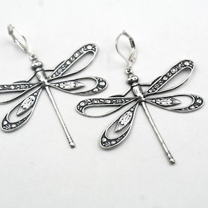 May include: A pair of silver-toned dragonfly earrings. Each earring features a detailed dragonfly design with openwork wings and small decorative accents. The earrings have a lever-back closure and are displayed against a white background.