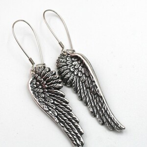 May include: A pair of silver-colored earrings shaped like angel wings. The earrings have a detailed feather design and long, thin hooks for pierced ears. The wings are approximately 2 inches long.