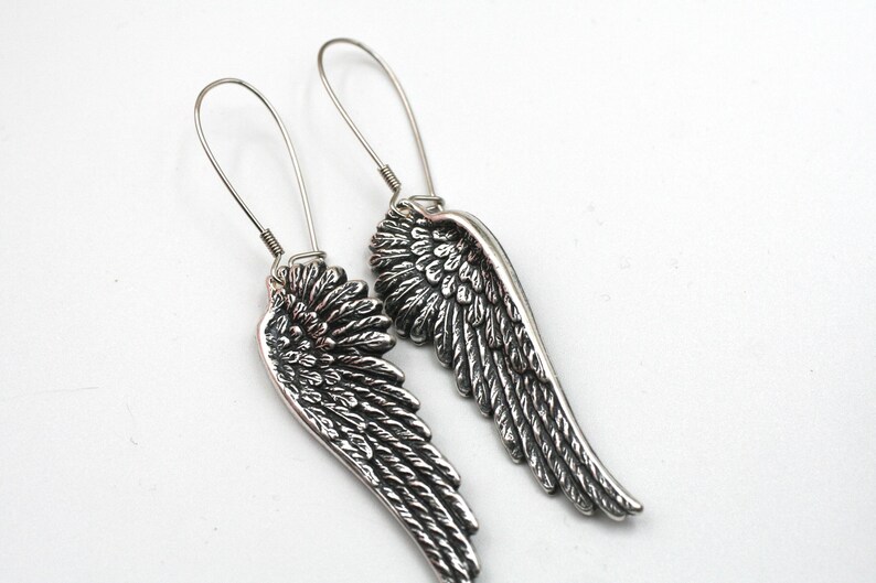 May include: A pair of silver-colored earrings shaped like angel wings. The earrings have a detailed feather design and long, thin hooks for pierced ears. The wings are approximately 2 inches long.