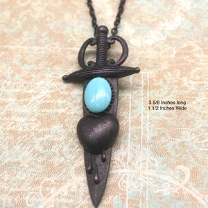 May include: A dark metal pendant necklace in the shape of a dagger, featuring a turquoise oval stone and a heart. The dagger is 3 5/8 inches long and 1 1/2 inches wide. The necklace has a dark chain.