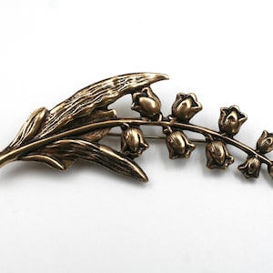 May include: A bronze-toned metal brooch shaped like a lily of the valley plant. The design features a textured, elongated leaf extending to the left, with a curved stem on the right supporting a series of small, bell-shaped flower blossoms.