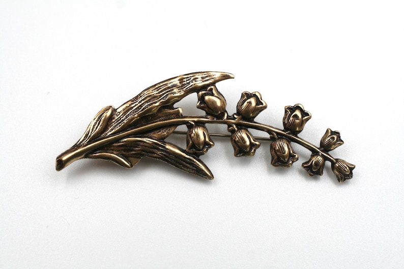 May include: A bronze-toned metal brooch shaped like a lily of the valley plant. The design features a textured, elongated leaf extending to the left, with a curved stem on the right supporting a series of small, bell-shaped flower blossoms.