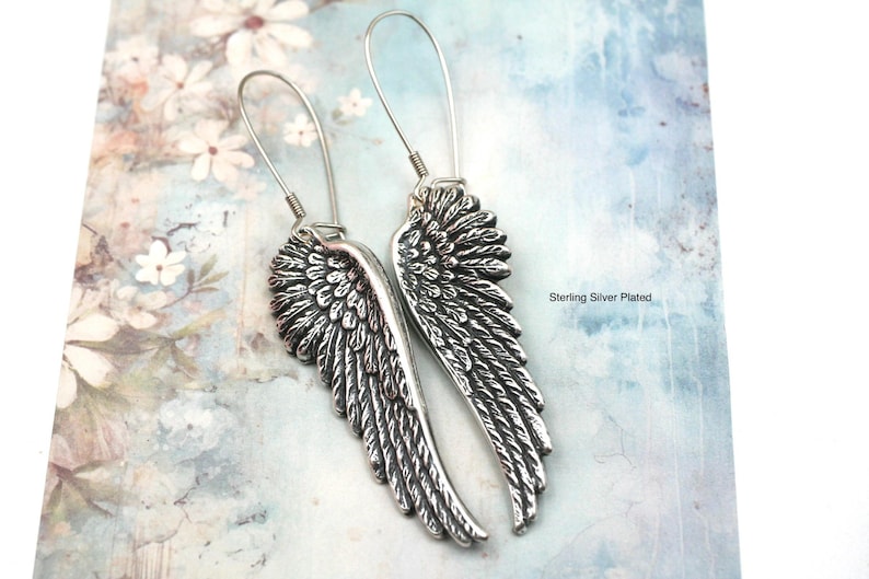May include: A pair of silver-colored wing earrings. The earrings have a detailed feather design and long, curved ear wires. The background is a light blue and white floral pattern. The text "Sterling Silver Plated" is visible.