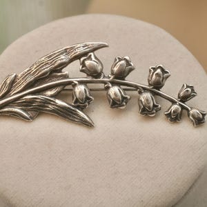 May include: A silver-toned brooch in the shape of a lily of the valley. The brooch features a detailed leaf and a stem with several bell-shaped flowers. The design is set against a neutral background, highlighting the intricate details of the jewelry.