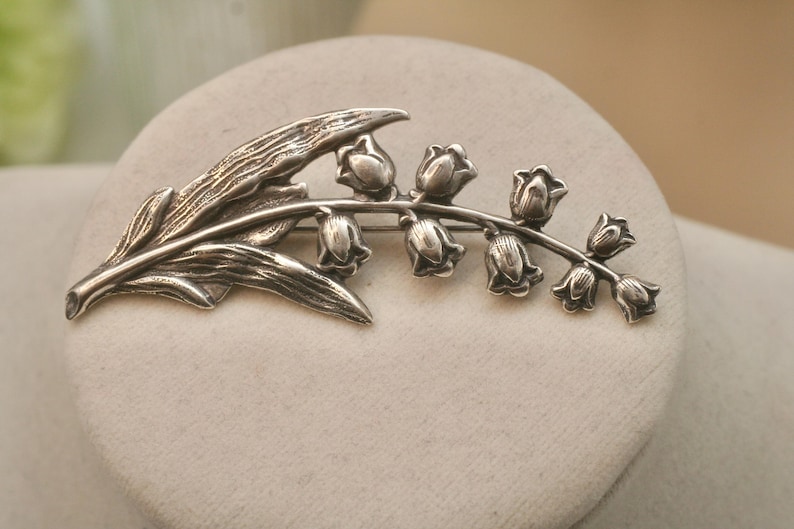 May include: A silver-toned brooch in the shape of a lily of the valley. The brooch features a detailed leaf and a stem with several bell-shaped flowers. The design is set against a neutral background, highlighting the intricate details of the jewelry.