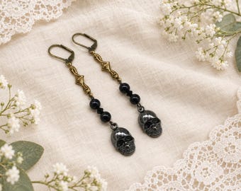 May include: A pair of dangle earrings featuring antique brass lever-back hooks, decorative metal links, two round black beads, and a dark metallic skull charm at the base of each earring. The design is symmetrical with a dangling drop construction.