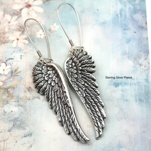 May include: A pair of silver-colored wing earrings. The earrings have a detailed feather design and long, curved ear wires. The background is a light blue and white floral pattern. The text "Sterling Silver Plated" is visible.