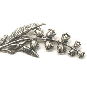 May include: A silver-toned brooch in the shape of a lily of the valley plant. The brooch features detailed leaves and bell-shaped flowers. The design is a botanical theme, suitable for adding a touch of nature-inspired elegance to any outfit.