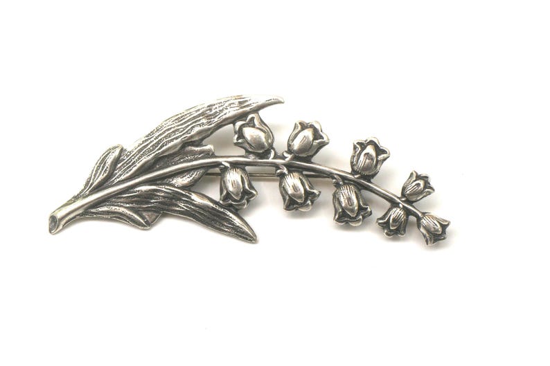 May include: A silver-toned brooch in the shape of a lily of the valley plant. The brooch features detailed leaves and bell-shaped flowers. The design is a botanical theme, suitable for adding a touch of nature-inspired elegance to any outfit.