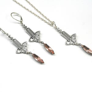 Op de afbeelding: Een zilveren ketting en oorbellen set met een delicaat, versierd ontwerp en hangende roze kristallen druppels.