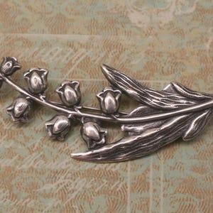 May include: A silver-toned brooch in the shape of a lily of the valley branch. The brooch features detailed leaves and bell-shaped flowers, with a textured finish. The design is a nature-inspired accessory.