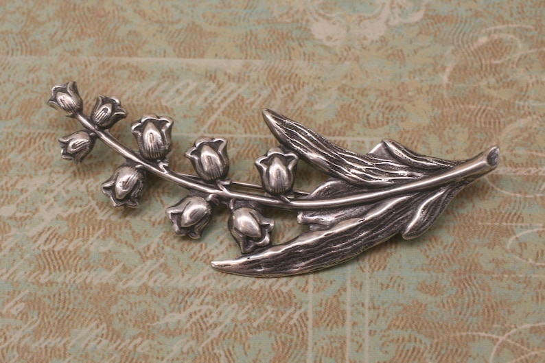 May include: A silver-toned brooch in the shape of a lily of the valley branch. The brooch features detailed leaves and bell-shaped flowers, with a textured finish. The design is a nature-inspired accessory.