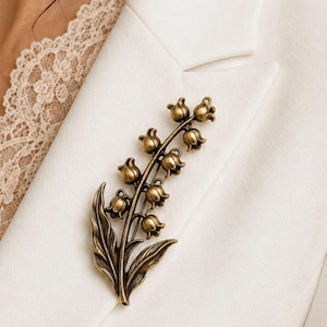 May include: A metallic bronze-toned brooch shaped like a lily of the valley flower stalk. The piece features multiple small, bell-shaped blossoms arranged along a central stem, accompanied by two elongated, pointed leaves at the base of the design.