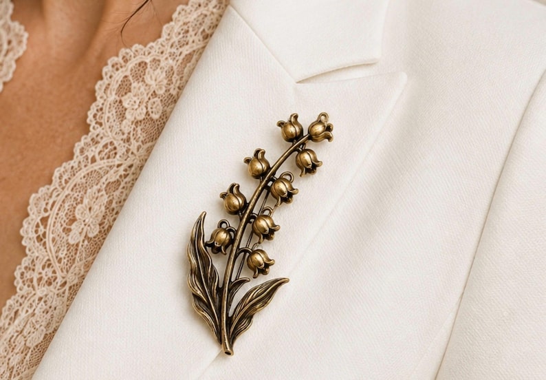 May include: A metallic bronze-toned brooch shaped like a lily of the valley flower stalk. The piece features multiple small, bell-shaped blossoms arranged along a central stem, accompanied by two elongated, pointed leaves at the base of the design.