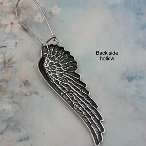 May include: A silver-toned wing-shaped earring with intricate feather detailing. The earring hangs from a simple wire hook. The back of the earring is hollow, as indicated by the text. The background is a soft, floral-patterned surface.
