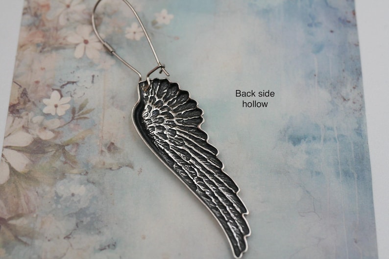 May include: A silver-toned wing-shaped earring with intricate feather detailing. The earring hangs from a simple wire hook. The back of the earring is hollow, as indicated by the text. The background is a soft, floral-patterned surface.