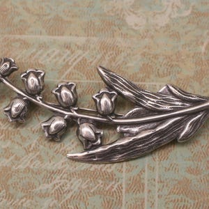 May include: A silver-toned brooch in the shape of a lily of the valley branch. The brooch features detailed leaves and bell-shaped flowers. The metal has a slightly aged appearance, suggesting a vintage or antique style. The brooch is a decorative accessory.