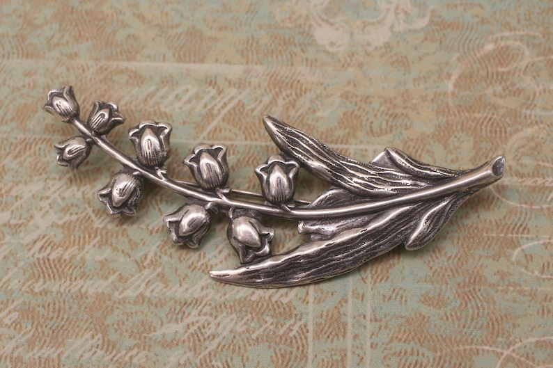 May include: A silver-toned brooch in the shape of a lily of the valley branch. The brooch features detailed leaves and bell-shaped flowers. The metal has a slightly aged appearance, suggesting a vintage or antique style. The brooch is a decorative accessory.